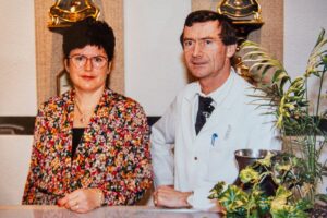 Portrait du couple fondateur de Gil Bonnet, derrière un comptoir, entouré d’horloges anciennes.
