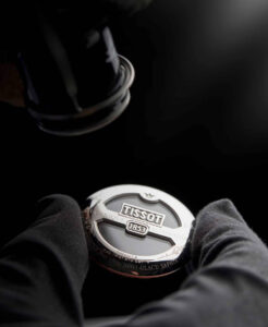 Technicien inspectant un fond de boîte Tissot avec une loupe, gants noirs et éclairage focalisé