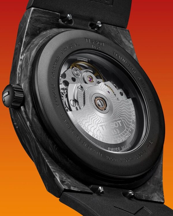 Vue rapprochée du fond de la montre Tissot PRX Powermatic 80 Carbone, montrant le mouvement automatique à travers le verre saphir et le boîtier noir en composite de carbone sur fond dégradé orange.