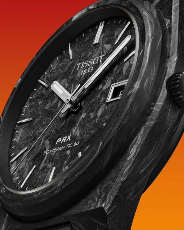 Gros plan sur la montre Tissot PRX Powermatic 80 Carbone, mettant en valeur le cadran noir en fibre de carbone, les aiguilles argentées et le boîtier texturé en composite de carbone sur fond orange.